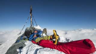 Ein Bergsteiger liegt auf dem schneebedeckten Gipfel eines Berges, umgeben von Ausrüstung und einem großen roten Schlafsack, mit klarem blauen Himmel und Wolken im Hintergrund.