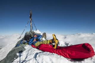 Ein Bergsteiger liegt auf dem schneebedeckten Gipfel eines Berges, umgeben von Ausrüstung und einem großen roten Schlafsack, mit klarem blauen Himmel und Wolken im Hintergrund.