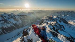Person in Daunenjacke sitzt mit Kamera und Schlafsack auf schneebedecktem Berggipfel bei Sonnenaufgang, umgeben von Bergpanorama.