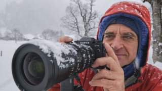Ein Mann in roter Winterjacke und blauer Mütze hält bei Schneefall eine schneebedeckte Kamera mit langem Objektiv. Im Hintergrund ist eine verschneite Landschaft mit Bäumen zu sehen.