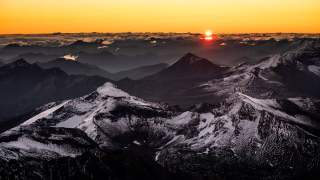 Berglandschaft mit schneebedeckten Gipfeln bei Sonnenaufgang und Wolken am Horizont. Die Sonne taucht die Szenerie in ein warmes, oranges Licht.