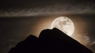 Vollmond leuchtet hinter einem dunklen Berggipfel mit einem kleinen Kreuz auf der Spitze, umgeben von leicht bewölktem Nachthimmel.