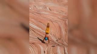 Ein Mensch mit blauer Mütze und gelber Hose macht einen Handstand auf einer rot-weißen, geschichteten Felsformation.