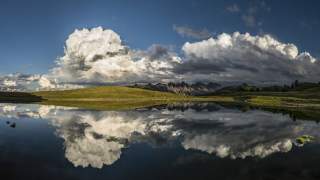Landschaft mit grünen Hügeln und Bergen, darüber große Wolken, die sich spiegelnd in einem ruhigen See darunter reflektieren.