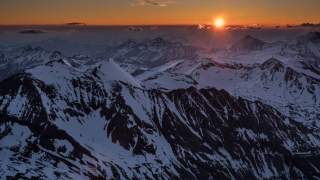 Schneebedeckte Berggipfel bei Sonnenuntergang mit orangefarbenem Himmel und vereinzelten Wolken.