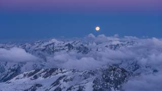Verschneite Berggipfel mit Wolken und voller Mond am Abendhimmel. Der Himmel zeigt einen Farbverlauf von Blau zu Lila.