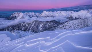 Verschneite Berglandschaft mit Nebelschwaden in den Tälern und einem violett-roten Himmel im Hintergrund.