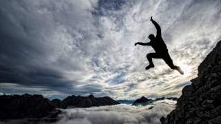 Silhouette einer Person, die von einem Felsen in luftiger Höhe vor bewölktem Himmel und Bergkulisse springt.