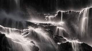 Wasserfälle fließen über dunkle, zerklüftete Felsen, wobei das Licht die Wasserbewegung in einem schwarz-weiß Bild betont. Die Szene wirkt dynamisch und kraftvoll.