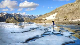 Eine Person steht auf einem teilweise zugefrorenen Bergsee, umgeben von schneebedeckten Bergen und felsigen Hängen unter einem blauen Himmel mit Wolken.