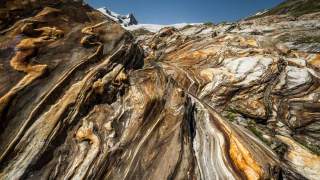 Nahaufnahme von farbig gemasertem Gestein mit verschlungenen, wellenförmigen Mustern und schneebedeckten Bergen im Hintergrund.