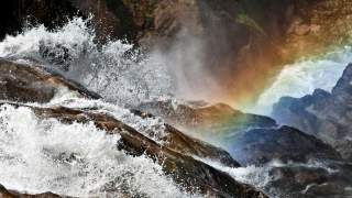 Wasser spritzt kraftvoll über Felsen, daneben ist ein Regenbogen, der sich durch den Wassernebel zieht.