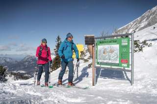 Zwei Skitourengeher in Winterkleidung schauen an einer beschilderten Informationsstation mit Karte und Hinweisen auf einer schneebedeckten Berglandschaft.