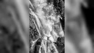 Eine Person macht einen Handstand auf einem Felsen vor einem großen, tosenden Wasserfall. Das Bild ist in Schwarz-Weiß gehalten.