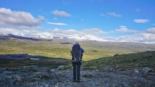 Eine Person mit großem Rucksack steht auf felsigem Untergrund und blickt auf eine weite, grüne Berglandschaft mit schneebedeckten Gipfeln unter blauem Himmel.