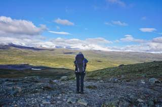 Eine Person mit großem Rucksack steht auf felsigem Untergrund und blickt auf eine weite, grüne Berglandschaft mit schneebedeckten Gipfeln unter blauem Himmel.