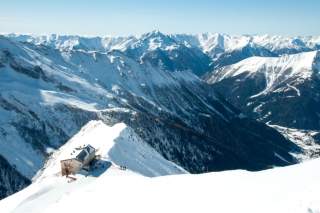 Schneebedeckte Alpenlandschaft mit einem Berghaus auf einem Gipfel und klar blauem Himmel. Im Hintergrund sind weitere schneebedeckte Berge und dunkle Waldtäler zu sehen.