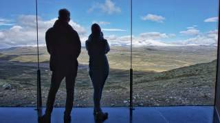Zwei Personen stehen vor einer großen Glasscheibe und blicken auf eine weite, gebirgige Landschaft mit blauem Himmel und Wolken.