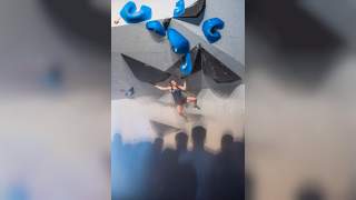 Eine Frau klettert an einer Boulderwand mit großen blauen und schwarzen Griffen, während mehrere Personen im Vordergrund zuschauen.