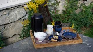 Tablett mit Kaffeekanne, Milchkrug, zwei blauen Tassen und Tellern sowie einem Stück Kuchen steht auf einem Gartentisch vor einem Stuhl und einer Steinmauer mit gelben Blumen.