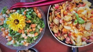 Bunte Gemüsepfanne in einer Pfanne neben einem frischen Salat mit einer gelben Blüte als Dekoration, daneben eine Schüssel mit Croutons.