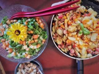 Bunte Gemüsepfanne in einer Pfanne neben einem frischen Salat mit einer gelben Blüte als Dekoration, daneben eine Schüssel mit Croutons.