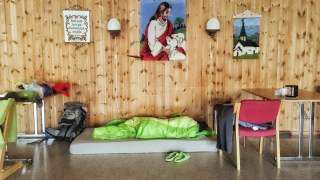 Eine Person liegt in einem grünen Schlafsack auf einer Matte auf dem Boden eines holzvertäfelten Raumes mit drei an der Wand hängenden Bildern. Daneben stehen Stühle, ein Tisch und Rucksäcke.