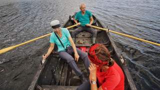 Drei Personen sitzen in einem Ruderboot auf einem Gewässer, wobei eine Person hinten das Boot mit zwei Rudern steuert.