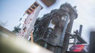 Zwei Personen klettern an einer mobilen Kletterwand vor einer großen Industrieanlage, im Vordergrund ist ein Banner mit der Aufschrift „DIE FINALS 2023 RHEIN-RUHR“ zu sehen.