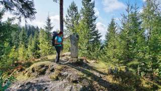 Eine Frau mit Rucksack und Wanderstock steht in einem bewaldeten Gebiet und berührt einen verzierten steinernen Wegweiser. Im Hintergrund sind Nadelbäume und ein bewölkter Himmel zu sehen.