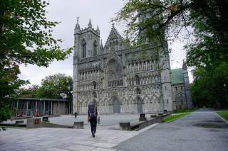 Ein Wanderer mit großem Rucksack geht auf eine große, verziere gotische Kirche zu, die von Bäumen eingerahmt ist. Der Himmel ist bewölkt.