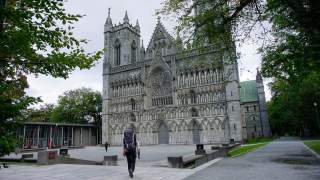 Ein Wanderer mit großem Rucksack geht auf eine große, verziere gotische Kirche zu, die von Bäumen eingerahmt ist. Der Himmel ist bewölkt.