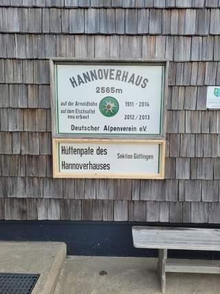 Schild am Hannnoverhaus auf 2565 Metern mit Informationen zur Höhe, den Baujahren und dem Deutschen Alpenverein e.V., befestigt an einer Holzschindelwand neben einer Bank.