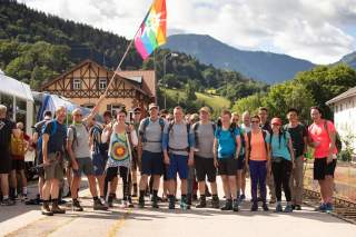 Eine Gruppe von Wanderern steht gemeinsam an einem Bahnhof mit Bergen im Hintergrund, eine Person hält eine bunte Flagge. Die Gruppe wirkt freundlich und bereit für eine Wanderung.