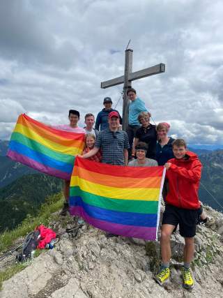 Eine Gruppe von Menschen steht auf einem Berggipfel vor einem Holzkreuz und hält zwei Regenbogenfahnen. Im Hintergrund sind bewaldete Berge und ein wolkiger Himmel zu sehen.
