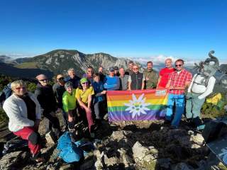Eine Gruppe von Wanderern posiert auf einem Berggipfel mit einer Regenbogenfahne des Deutschen Alpenvereins, im Hintergrund sind Berge und blauer Himmel zu sehen.