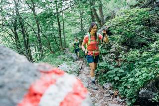 Zwei Wanderer mit Rucksäcken und Wanderstöcken gehen einen felsigen Waldweg entlang, im Vordergrund ist ein Felsen mit roter und weißer Markierung zu sehen.