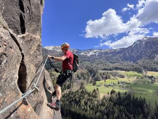 Ein Klettersteiggeher mit Helm und Kletterausrüstung sichert sich an einem Drahtseil an einer steilen Felswand, im Hintergrund Wiesen, Wälder und schneebedeckte Berge unter blauem Himmel mit Wolken.