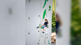 Ein Mädchen klettert gesichert an einer künstlichen Kletterwand mit grünen Griffen und Tritten.