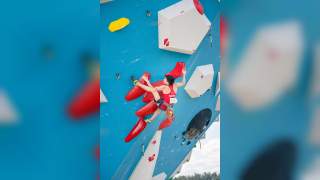 Ein junger Kletterer in rotem Outfit klettert an einer blauen Kletterwand mit roten und weißen Griffen, gesichert mit einem Klettergurt.