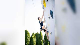 Junger Mann klettert an einer künstlichen Kletterwand im Freien, umgeben von hohen Bäumen.
