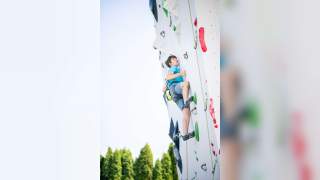 Ein junger Mann klettert an einer künstlichen Kletterwand im Freien, trägt Kletterausrüstung und blickt konzentriert nach oben. Im Hintergrund sind grüne Bäume und ein klarer Himmel zu sehen.