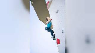 Junge Frau klettert an einer Kletterwand, gesichert mit Klettergurt und Seil, vor blauem Himmel.