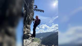 Ein Klettersteiggeher mit Helm und Ausrüstung steht an einer Felswand, im Hintergrund Berge, Wälder und ein Tal; über dem Mann ein Metallzeichen mit der Aufschrift „Ratschings Racines“ und einem Steinbock.