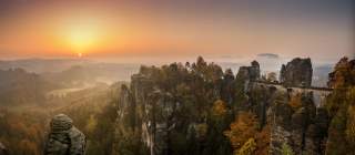 Sonnenaufgang über einem herbstlichen Felsmassiv mit einer Steinbrücke, umgeben von buntem Wald und nebligen Tälern.