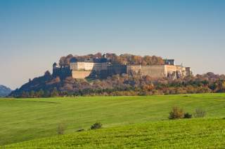 Blick auf eine große Burg auf einem bewaldeten Hügel mit herbstlich gefärbten Bäumen, davor eine weite grüne Wiese unter klarem Himmel.