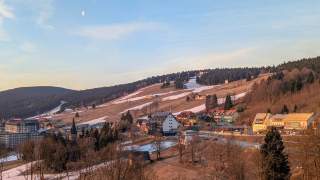 Ein sanfter Berghang mit einer Siedlung im Vordergrund und einem Skilift im Hintergrund. Es liegt Restschnee.