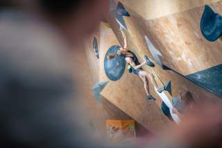 Eine Frau klettert an einer Indoor-Boulderwand und greift nach einem Griff, während sie in sportlicher Kleidung aktiv ist. Im Vordergrund ist unscharf ein weiterer Mensch zu sehen.