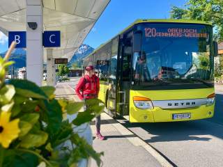 Eine Person mit rotem Rucksack steht an einer Bushaltestelle neben einem gelben Linienbus mit der Zielanzeige „120 Oberjoch Einhaus-Weißensee“, im Hintergrund Berge und blauer Himmel.