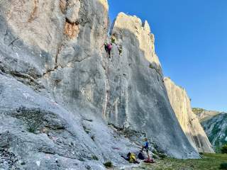 Eine Person klettert an einer steilen Felswand, während eine zweite Person mit Kletterausrüstung unten sichert, umgeben von einer felsigen Landschaft unter blauem Himmel.